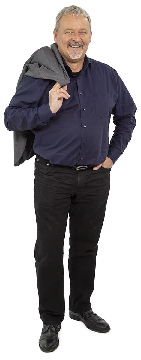 Lächelnder Mann in dunklem Hemd und Hose, der eine graue Jacke über der Schulter trägt, steht vor einem weißen Hintergrund.