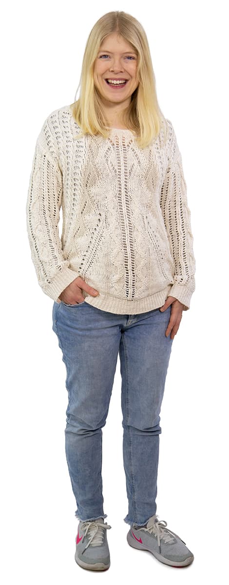 Lächelnde Person mit langen blonden Haaren in einem cremefarbenen Pullover, blauen Jeans und grauen Turnschuhen, die mit einer Hand in der Tasche steht.