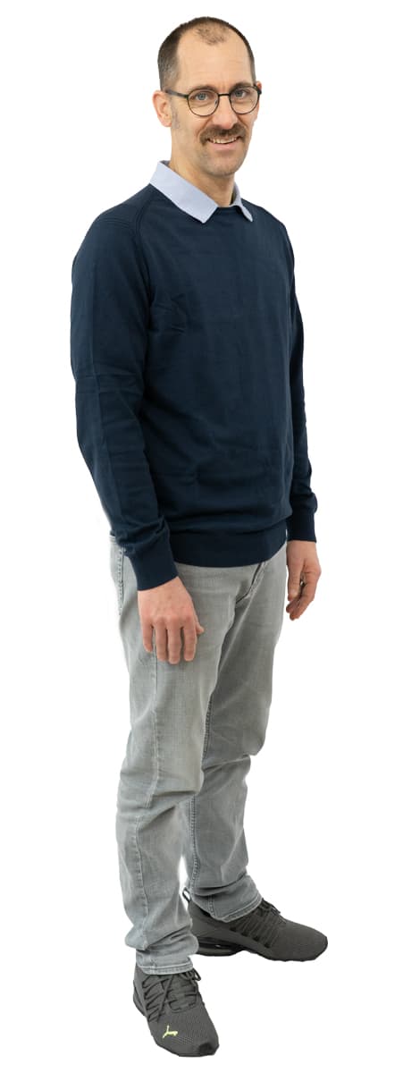 Mann mit Brille, Schnurrbart und kurzen Haaren, bekleidet mit einem blauen Pullover, hellen Jeans und grauen Turnschuhen, steht und lächelt.