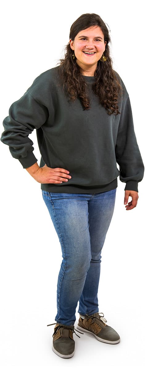 Frau mit langen lockigen Haaren in einem dunklen Sweatshirt, Jeans und Turnschuhen steht und lächelt vor einem weißen Hintergrund.