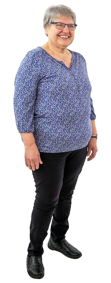 Lächelnde ältere Frau mit kurzen grauen Haaren, Brille, blau gemustertem Oberteil, schwarzer Hose und schwarzen Schuhen steht.