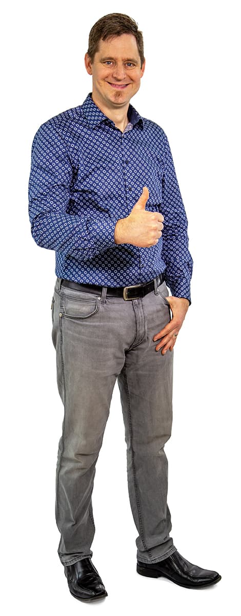 Ein Mann in einem blau gemusterten Hemd und grauen Jeans steht da, zeigt den Daumen nach oben und lächelt in die Kamera.