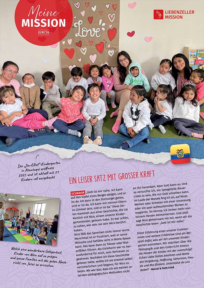 Eine Gruppe lächelnder Kinder und zwei Frauen sitzen und knien auf dem Boden eines Klassenzimmers, hinter ihnen sind Herzen an der Wand angebracht.