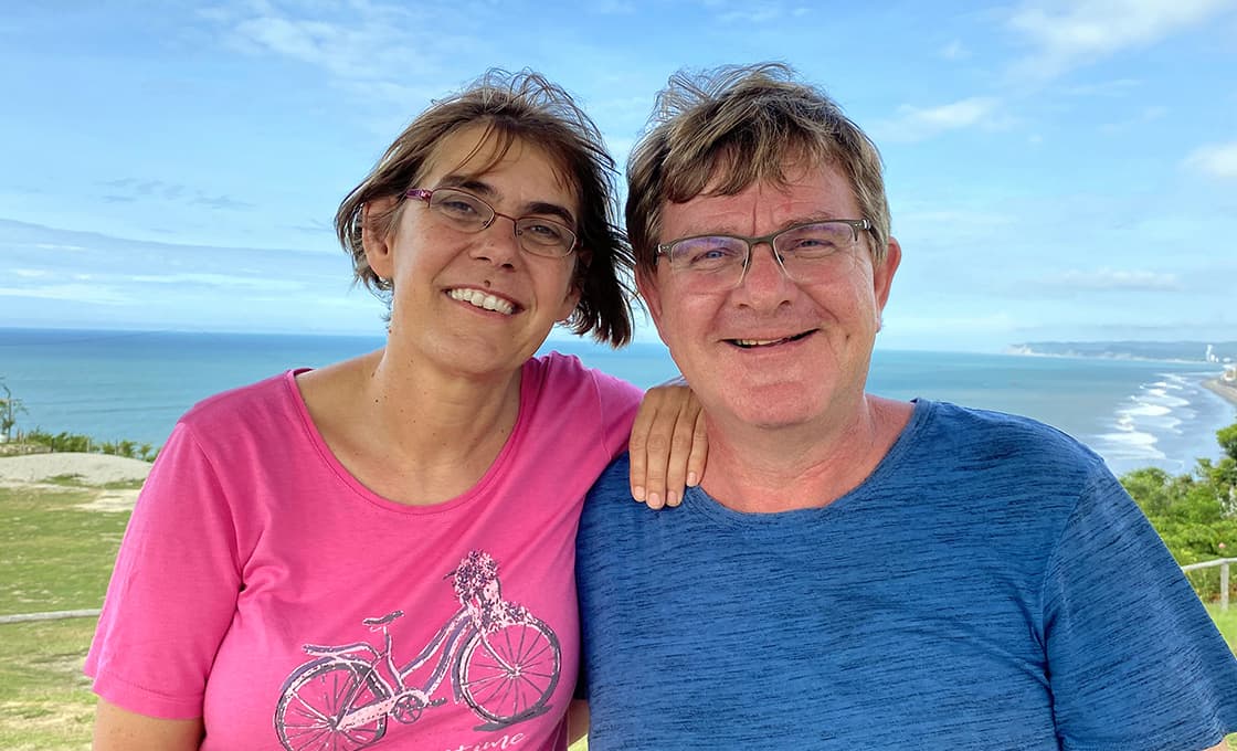 Lächelnde Frau und Mann mit Brille posieren zusammen im Freien mit Meer und blauem Himmel im Hintergrund.