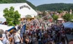 Eine große Menschenmenge versammelt sich bei einem Freiluftfestival in einer kleinen Stadt, die von grünen Hügeln und Gebäuden umgeben ist.