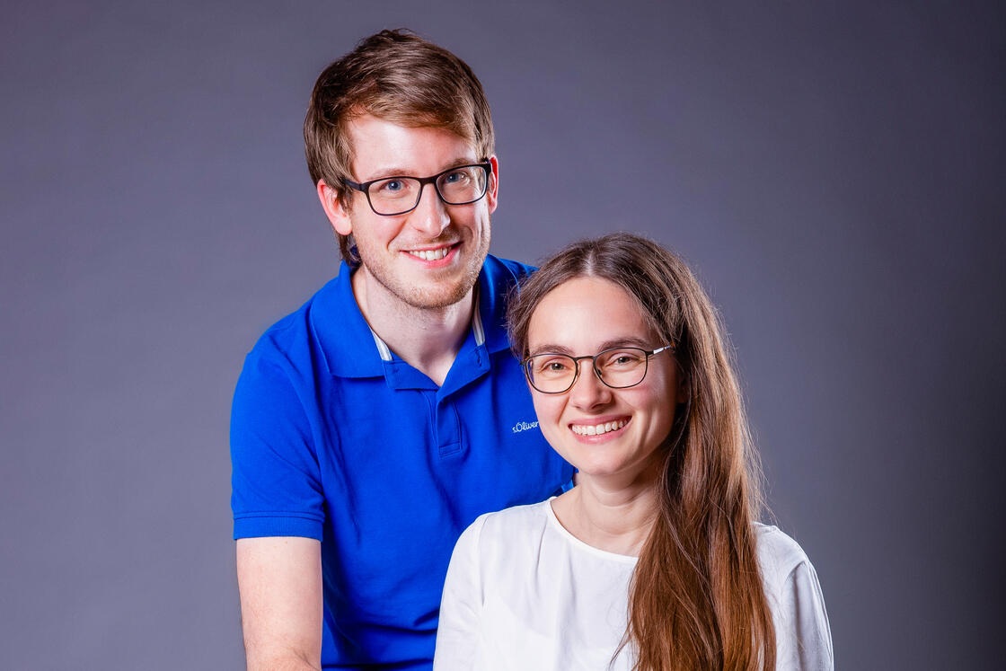 Ein lächelnder Mann in einem blauen Polohemd steht neben einer Frau in einem weißen Oberteil vor einem schlichten grauen Hintergrund.
