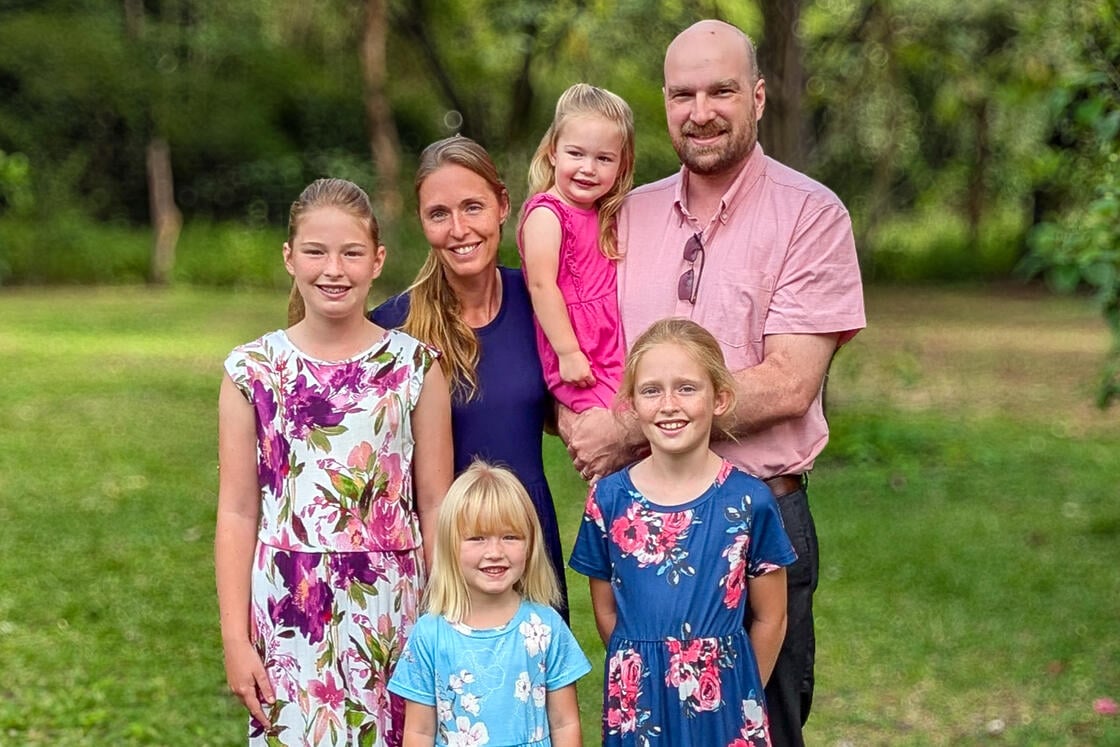 Eine fünfköpfige Familie posiert im Freien – drei kleine Mädchen und zwei Erwachsene, die alle in einer grünen, bewaldeten Umgebung lächeln.