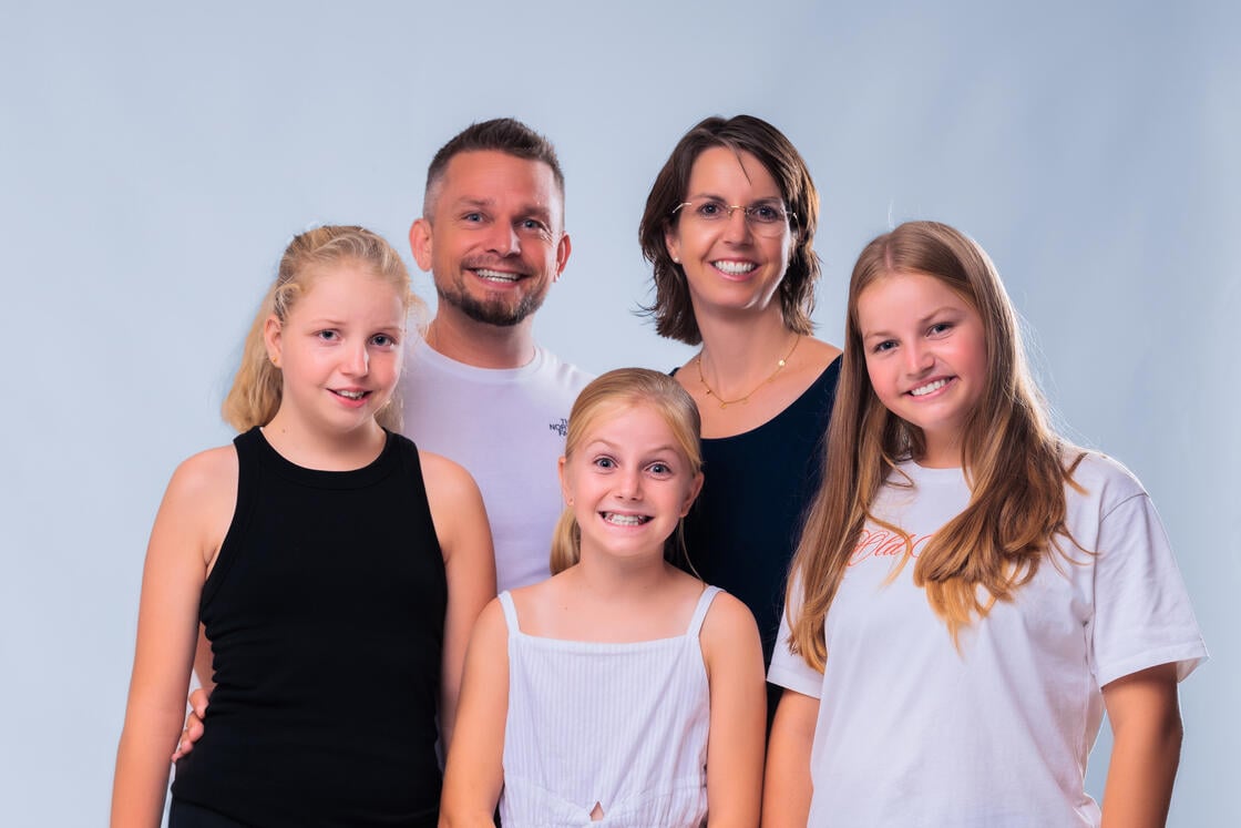Eine lächelnde fünfköpfige Familie – zwei Erwachsene und drei kleine Mädchen – posiert gemeinsam vor einem hellblauen Hintergrund.