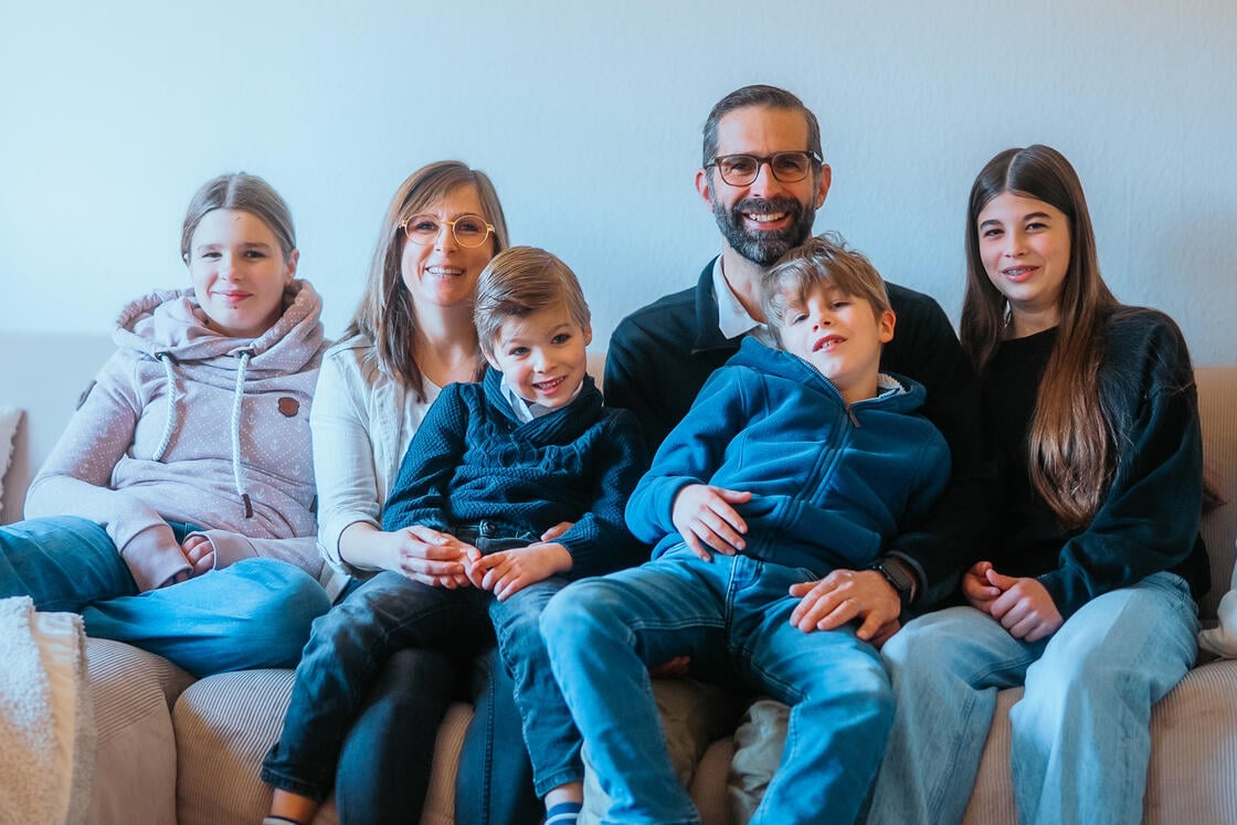 Eine lächelnde sechsköpfige Familie sitzt gemeinsam auf einem Sofa – die Eltern und ihre vier Kinder – in einem Zimmer.