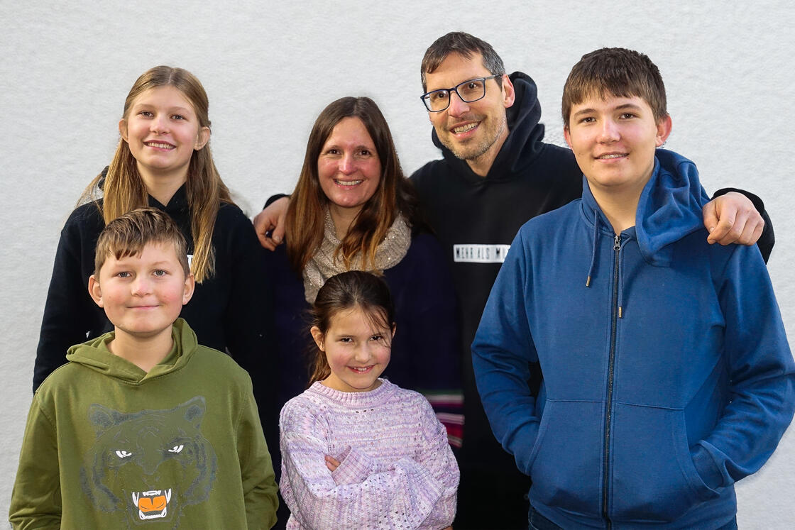 Eine sechsköpfige Familie, bestehend aus zwei Erwachsenen und vier Kindern, posiert gemeinsam und lächelt vor einem schlichten, hellen Hintergrund.