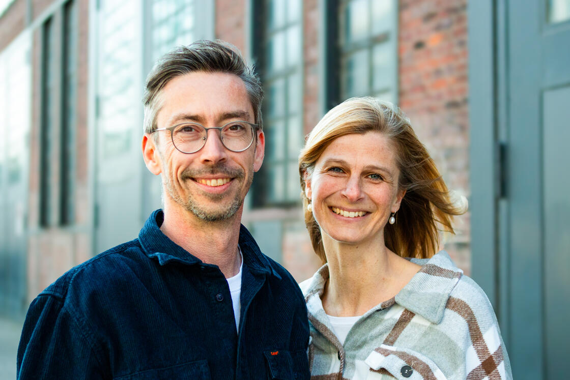 Ein lächelnder Mann und eine lächelnde Frau stehen gemeinsam im Freien vor einem Backsteingebäude.