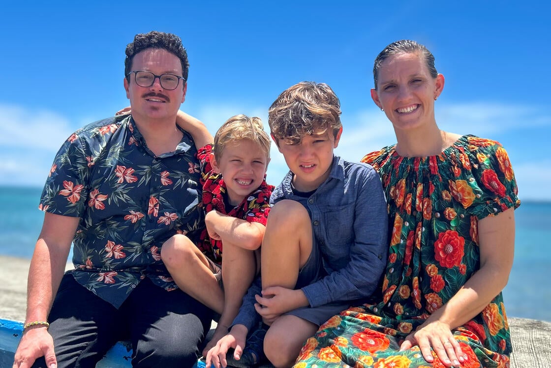 Eine lächelnde vierköpfige Familie posiert gemeinsam im Freien am Meer; alle tragen bunte Kleidung mit Blumenmustern.