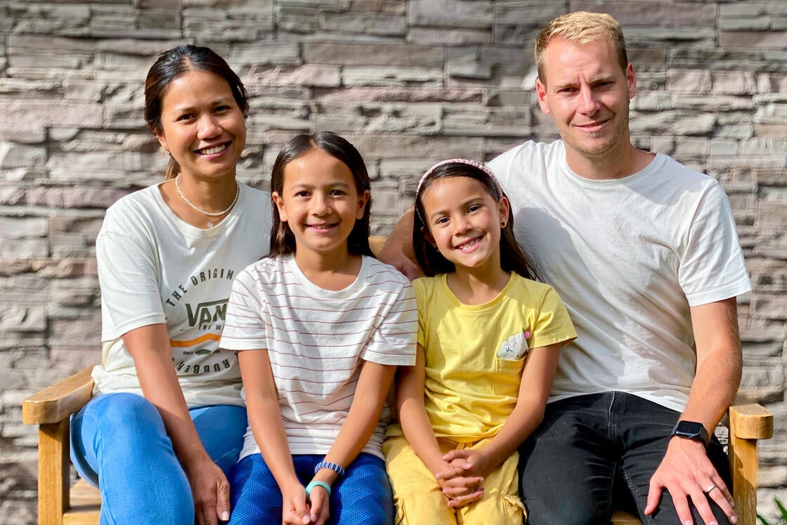 Eine lächelnde vierköpfige Familie sitzt auf einer Bank vor einer Steinmauer; dazu gehören zwei Erwachsene und zwei kleine Kinder.