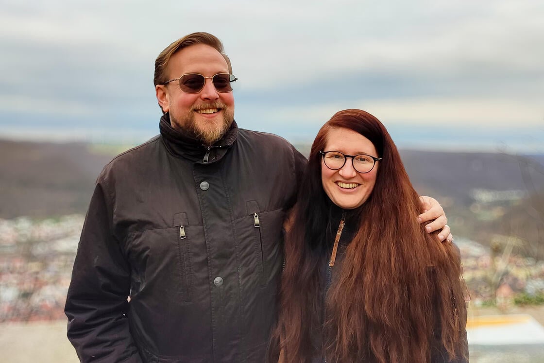 Ein lächelnder Mann und eine lächelnde Frau mit Brille posieren im Freien, im Hintergrund sind Hügel und ein bewölkter Himmel zu sehen.