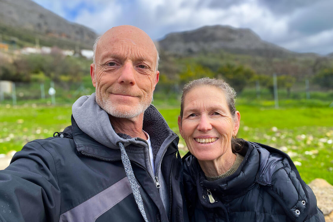 Ein Mann und eine Frau in Jacken, die im Freien lächeln, im Hintergrund grünes Gras und Berge.
