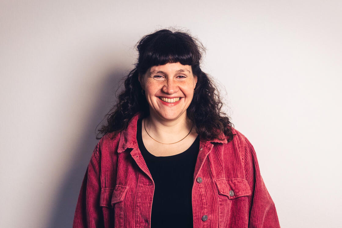 Eine Frau mit lockigem braunem Haar, die lächelt, trägt eine rosa Cordjacke über einem schwarzen Shirt und steht vor einem hellen Hintergrund.