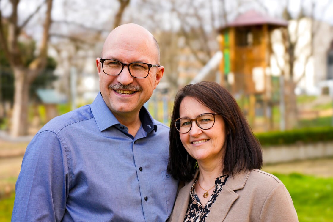 Ein lächelnder Mann und eine lächelnde Frau mit Brille stehen gemeinsam im Freien in einem Park; im Hintergrund sind Bäume und ein Spielplatz verschwommen zu sehen.