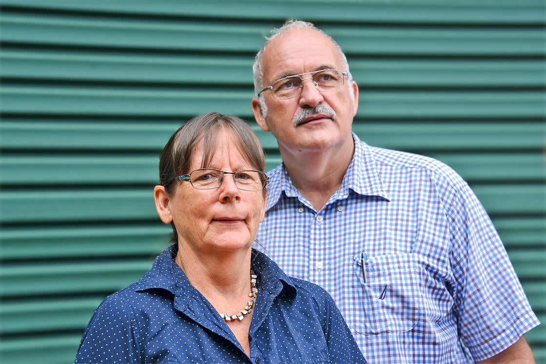 Zwei ältere Menschen stehen vor einer grün gestreiften Wand; beide tragen eine Brille und blau gemusterte Hemden.