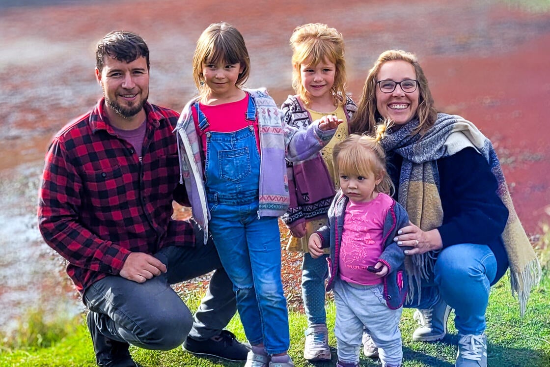 Eine lächelnde fünfköpfige Familie posiert im Freien auf einer Wiese in der Nähe eines Sees – zwei Erwachsene und drei kleine Kinder.