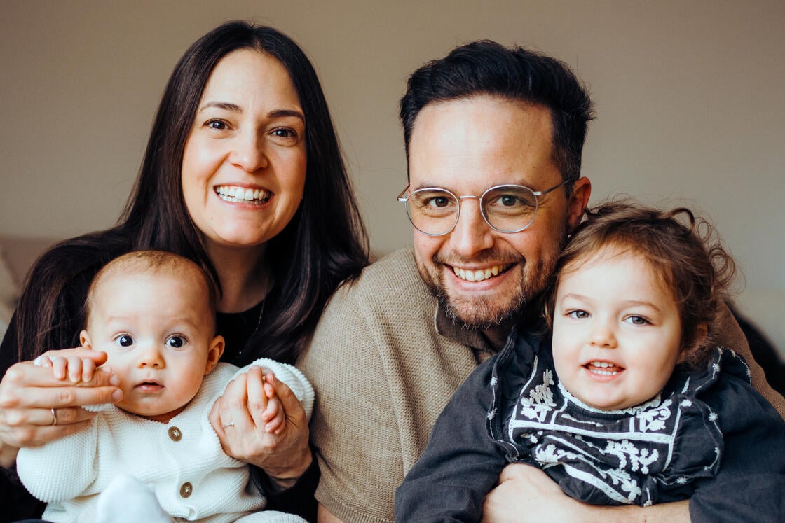 Eine lächelnde vierköpfige Familie posiert gemeinsam in einem Raum, wobei die Eltern ein Baby und ein Kleinkind im Arm halten.