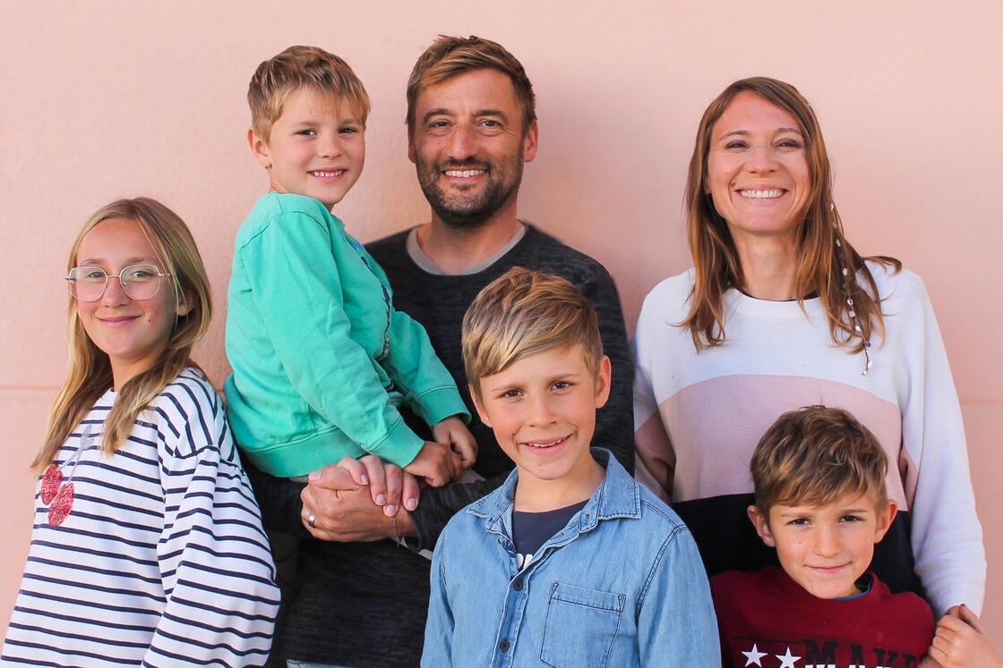 Eine fünfköpfige Familie – zwei Erwachsene und drei Kinder – lächelt gemeinsam vor einer hellrosa Wand.