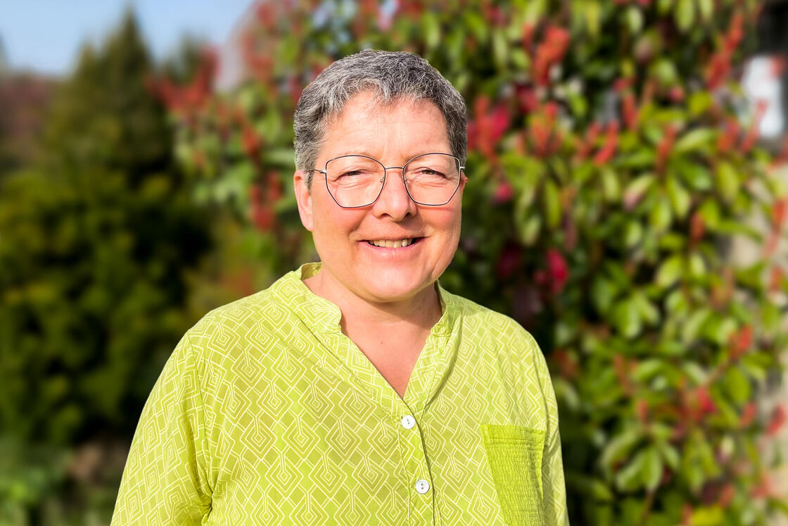 Eine lächelnde Person mit kurzen grauen Haaren und Brille, die ein gelbes Hemd trägt und draußen in einem Garten steht.