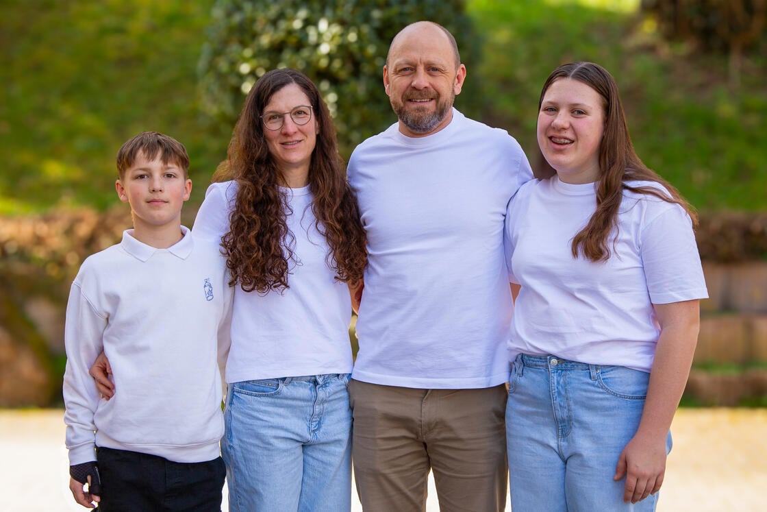 Eine vierköpfige Familie, alle in weißen Hemden und Jeans, steht an einem sonnigen Tag im Freien eng beieinander und lächelt.