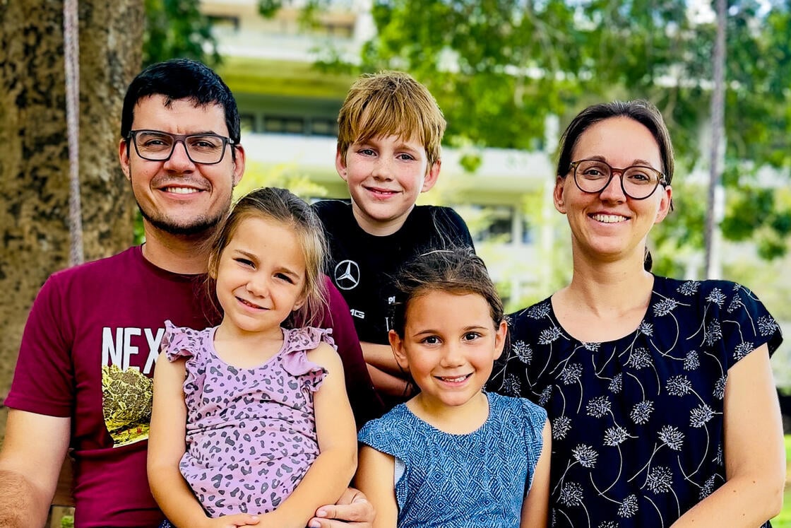Eine lächelnde fünfköpfige Familie – zwei Erwachsene und drei kleine Kinder – posiert im Freien vor Bäumen und einem Gebäude.