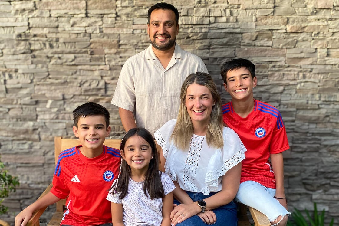 Eine lächelnde fünfköpfige Familie posiert im Freien; zwei Jungen tragen rote Fußballtrikots, ein Mädchen und die Erwachsenen stehen vor einer Steinmauer.