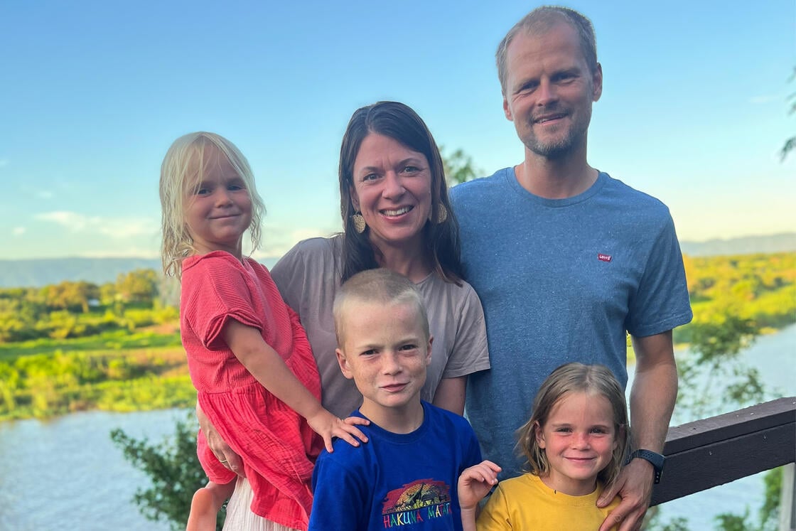 Eine lächelnde fünfköpfige Familie posiert im Freien am Ufer eines Flusses, im Hintergrund sind grüne Bäume und ein blauer Himmel zu sehen.