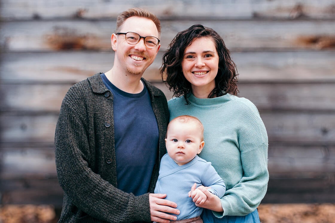 Ein lächelnder Mann und eine lächelnde Frau stehen draußen vor einer verwitterten Holzwand und halten ein Baby im Arm.