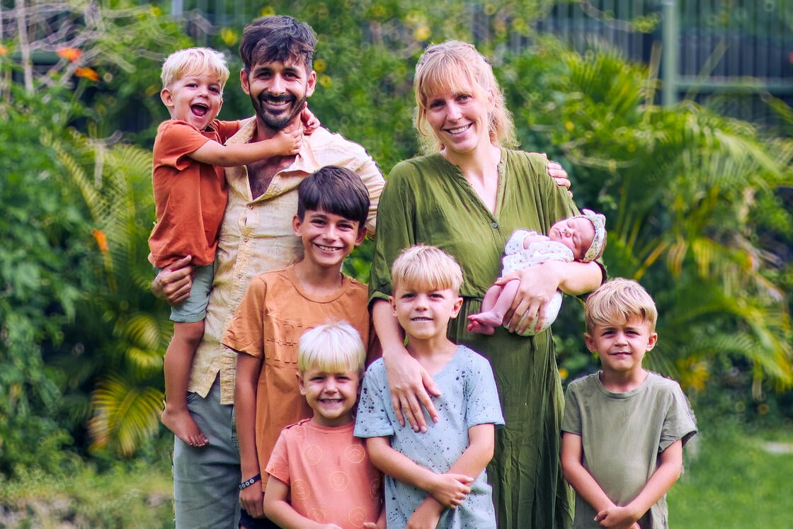Eine lächelnde Familie mit sechs kleinen Kindern posiert draußen inmitten von Grün; ein Baby wird von der Mutter im Arm gehalten.