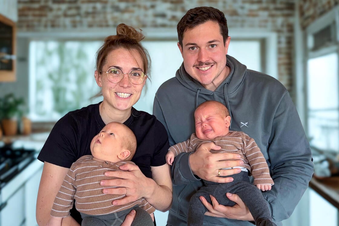 Ein lächelndes Paar hält in einer gemütlichen Küche zwei Babys in identischer Kleidung im Arm. Das eine Baby weint, das andere schaut ruhig nach oben.
