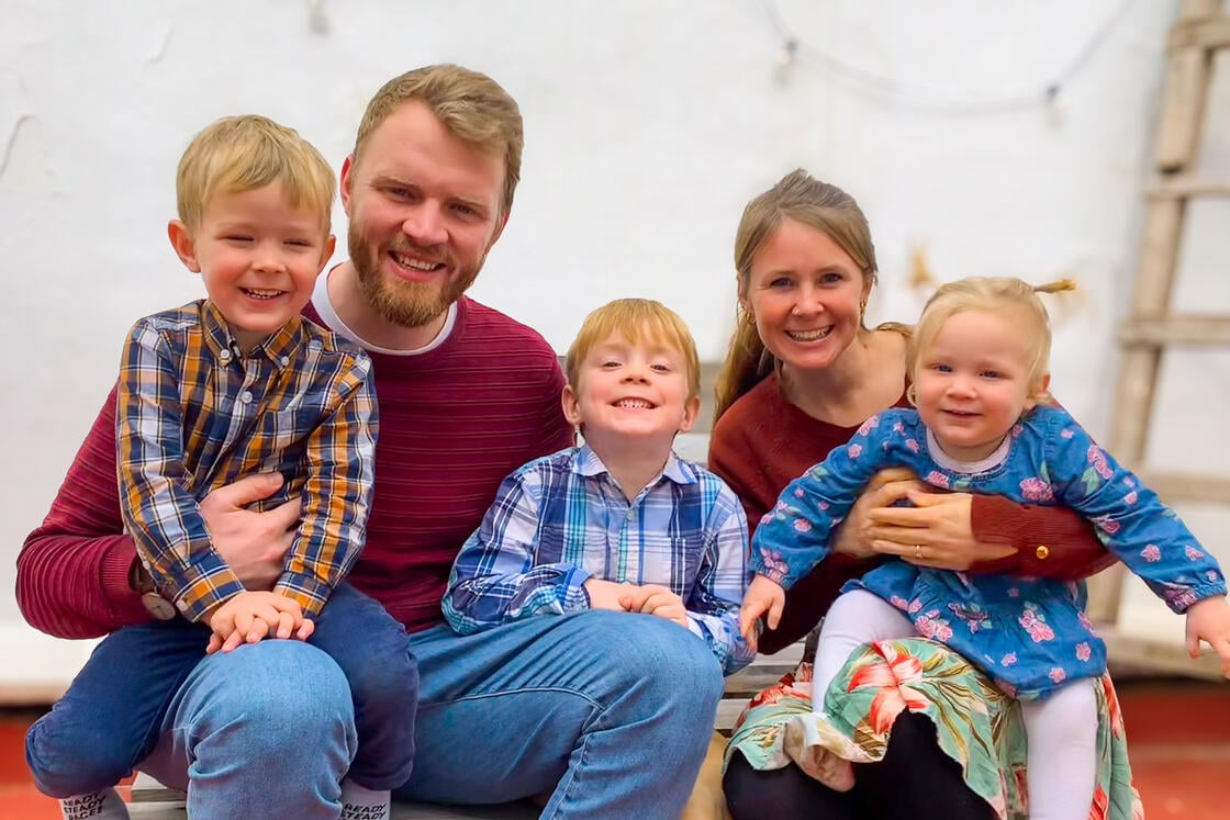Eine lächelnde fünfköpfige Familie – zwei Erwachsene und drei kleine Kinder – sitzt eng beieinander auf einer Bank in einem Raum.