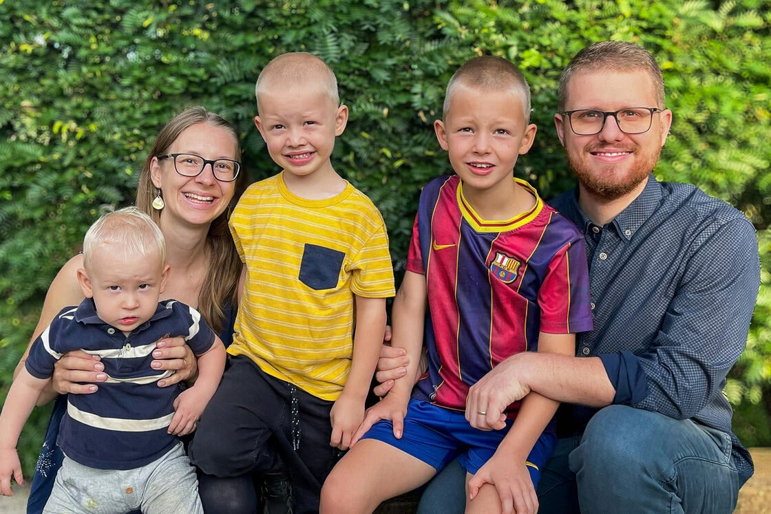 Eine lächelnde fünfköpfige Familie posiert im Freien vor dem Hintergrund von grünem Laub. Drei kleine Jungen sitzen neben ihren Eltern.
