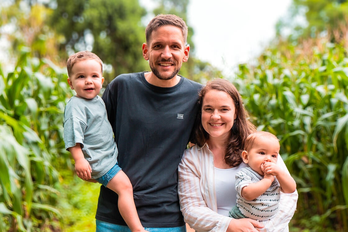 Eine lächelnde vierköpfige Familie steht draußen auf einer grünen, bewachsenen Wiese; die Eltern halten zwei kleine Kinder im Arm.
