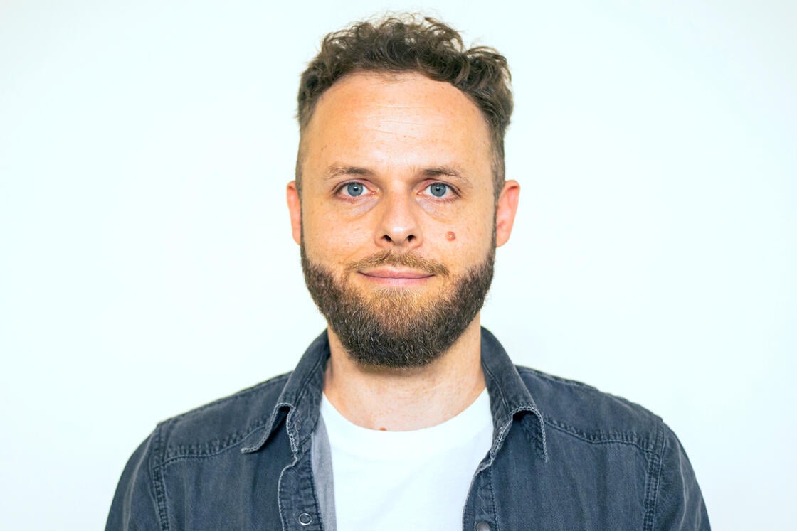 Ein Mann mit kurzen Locken und Bart, der ein Jeanshemd über einem weißen T-Shirt trägt und in die Kamera blickt.