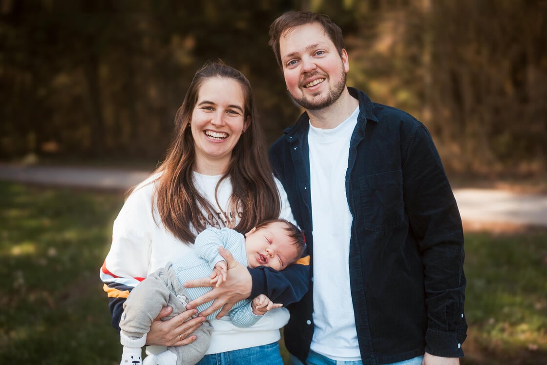 Ein lächelndes Paar steht im Freien; die Frau hält ein schlafendes Baby im Arm, im Hintergrund sind verschwommene Bäume zu sehen.