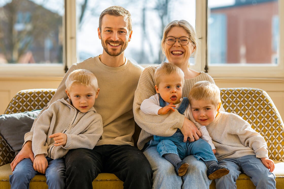 Ein lächelndes Paar sitzt mit drei kleinen Kindern auf einem Sofa in einem hellen, sonnendurchfluteten Raum.