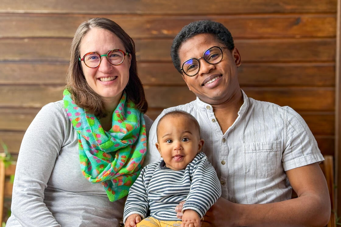 Ein lächelndes Paar mit Brille posiert in einem Raum und hält ein fröhliches Baby im Arm, das die Zunge herausstreckt.