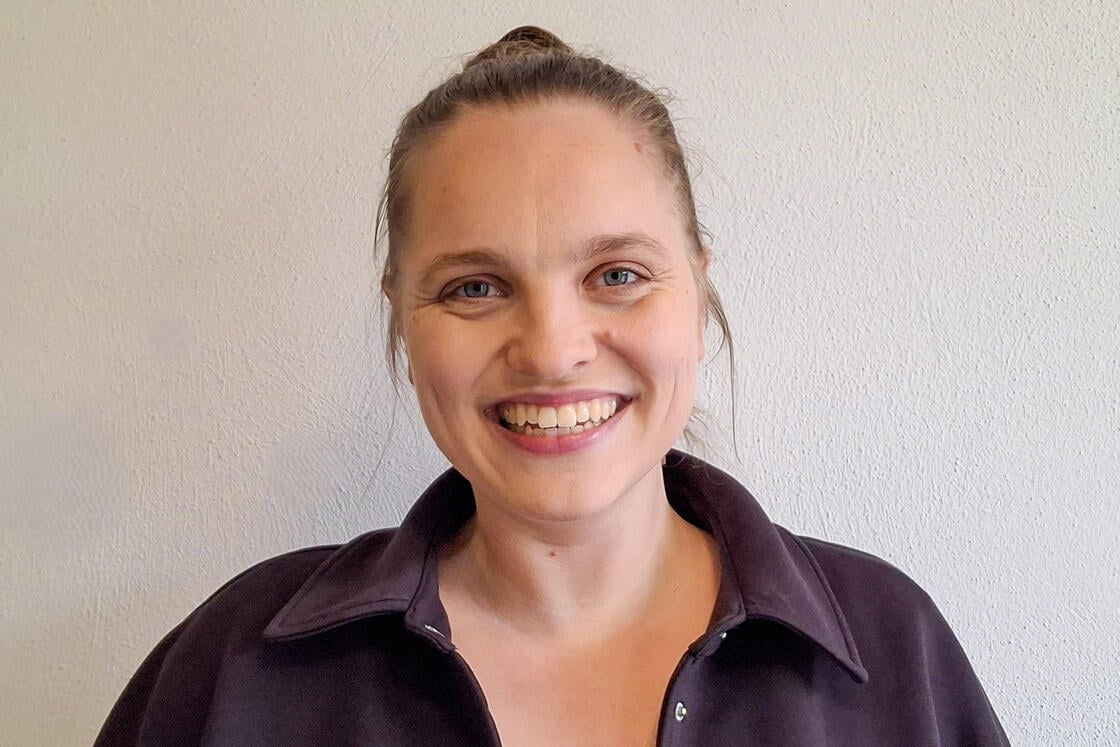 Eine lächelnde Person mit hellbraunen Haaren, die zu einem Dutt zusammengebunden sind, trägt ein dunkles Hemd mit Kragen und steht vor einer hellen Wand.