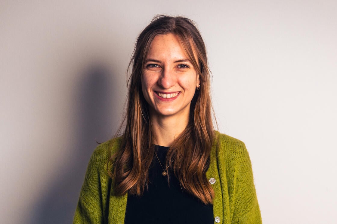 Eine Frau mit langen braunen Haaren, die einen grünen Pullover trägt und vor einer schlichten, hellen Wand lächelt.