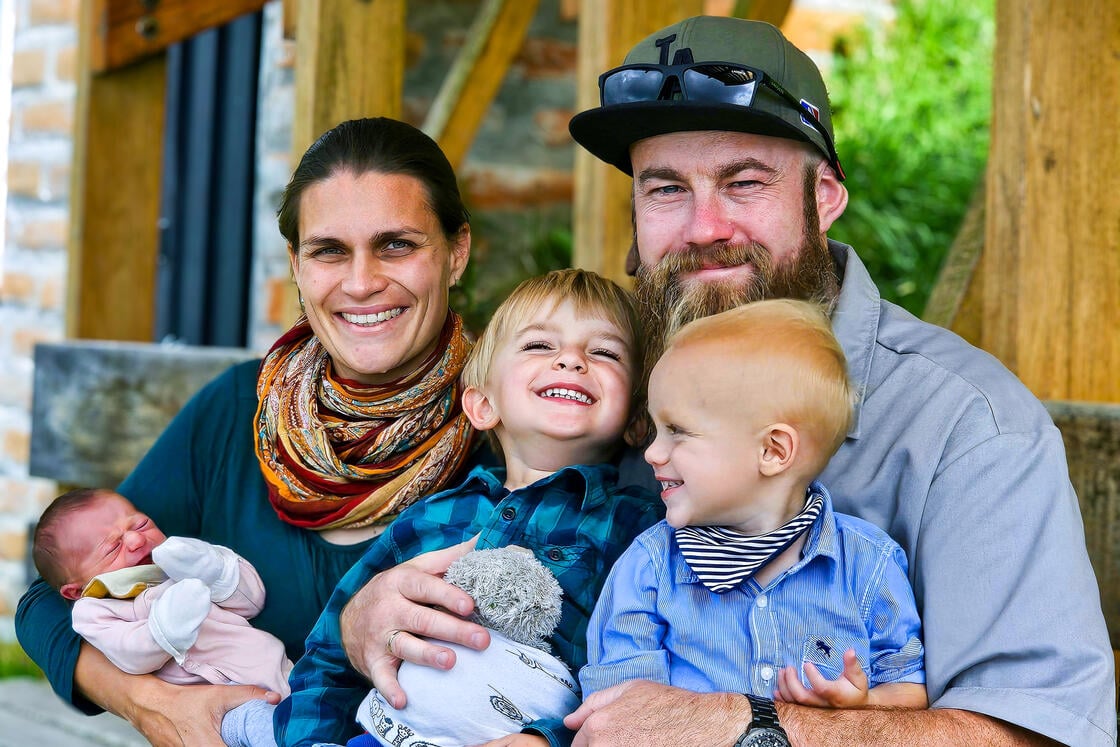 Eine lächelnde Familie mit zwei Erwachsenen und drei kleinen Kindern posiert gemeinsam im Freien; das jüngste Kind ist ein Baby in rosa Kleidung.