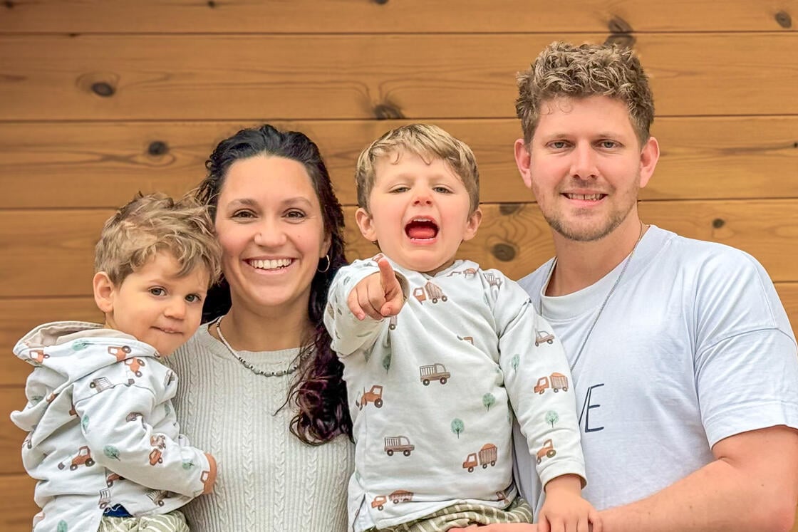 Eine lächelnde vierköpfige Familie posiert vor einer Holzwand; eines der Kinder zeigt aufgeregt in die Kamera.