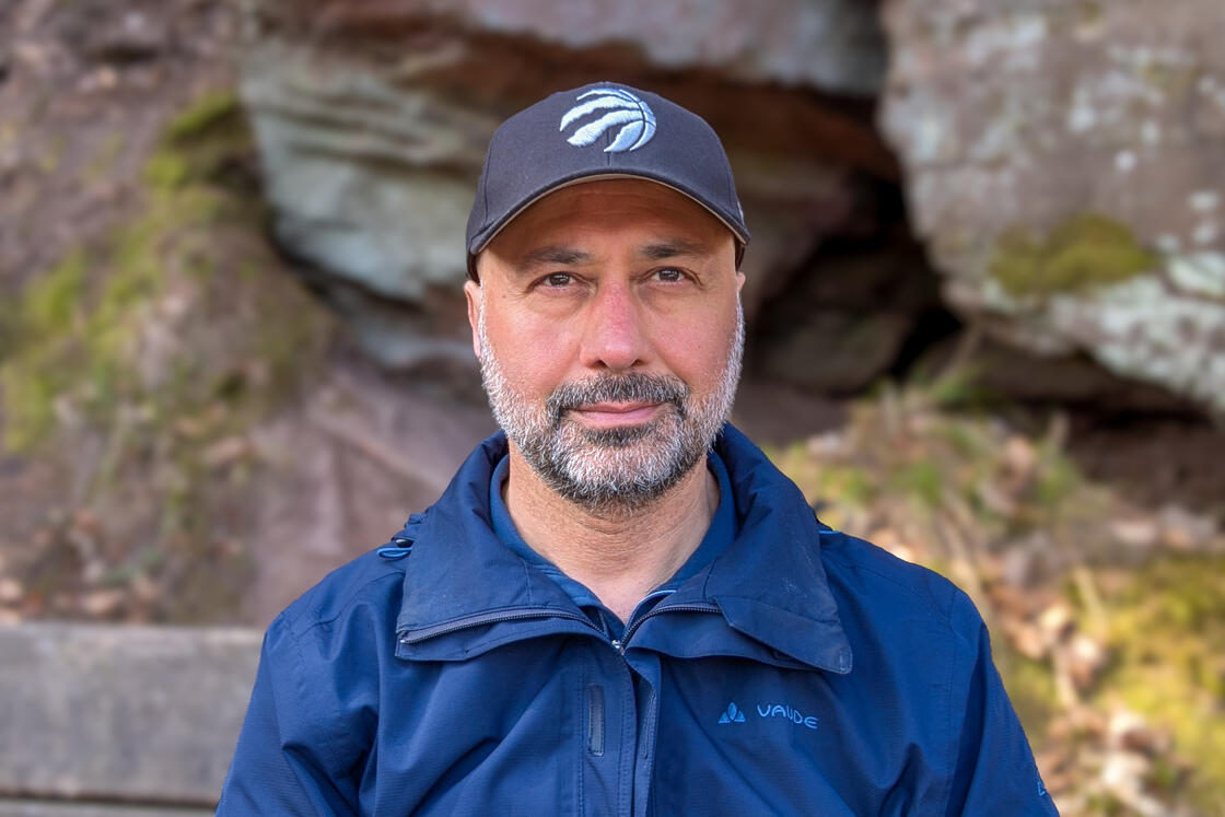 Ein Mann in einer blauen Jacke und einer schwarzen Mütze steht im Freien vor Felsen und Grünpflanzen und blickt in die Kamera.
