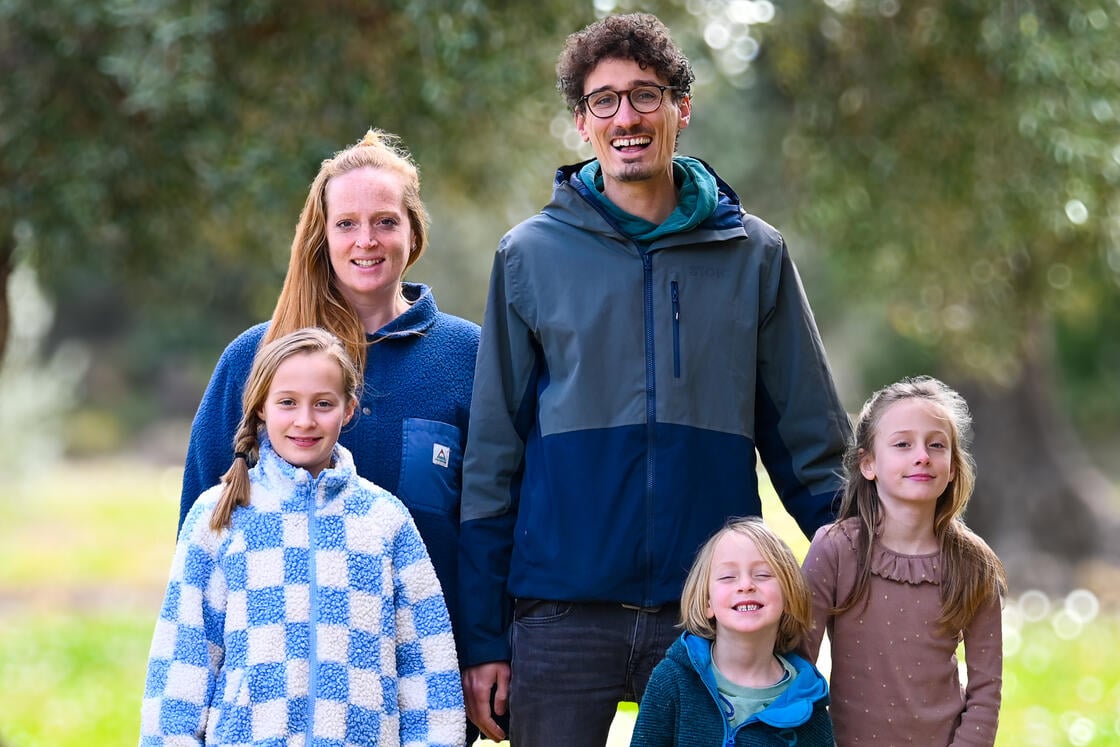 Eine lächelnde fünfköpfige Familie posiert im Freien: Zwei Erwachsene und drei Kinder stehen eng beieinander.