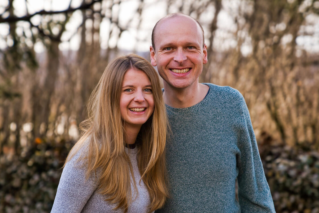 Ein lächelnder Mann und eine lächelnde Frau stehen eng beieinander im Freien, im verschwommenen Hintergrund sind Bäume und Blätter zu sehen.
