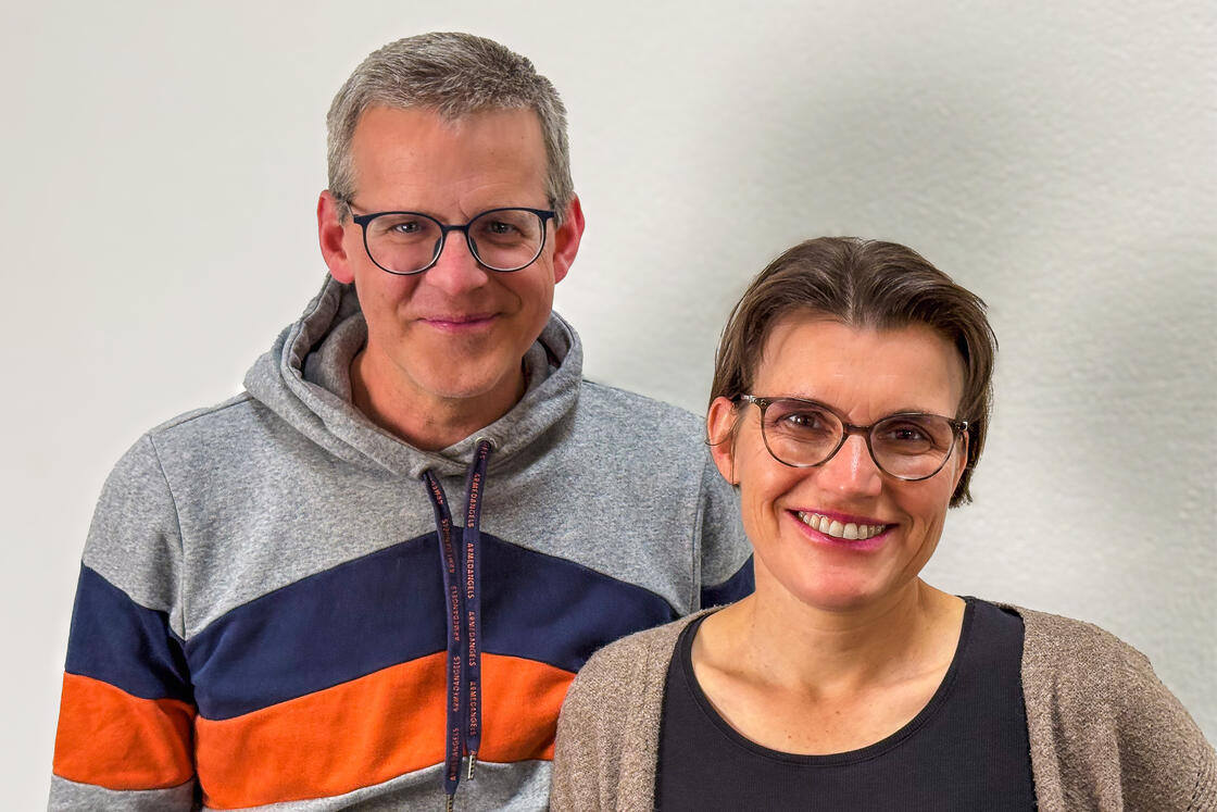 Ein Mann mit Brille und einem gestreiften Kapuzenpullover steht neben einer lächelnden Frau mit Brille und einem hellbraunen Pullover.