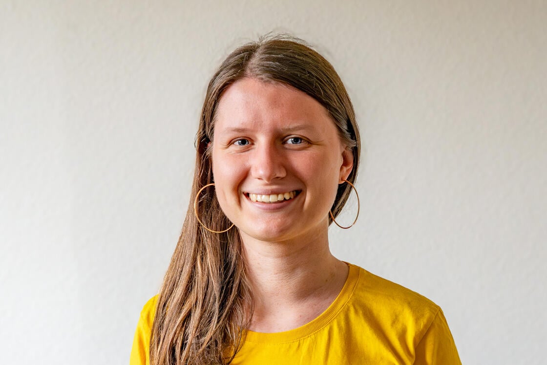 Eine Frau mit langen braunen Haaren, die ein gelbes Shirt und große Creolen trägt und in die Kamera lächelt.