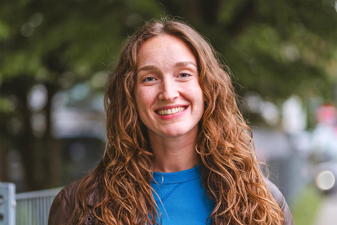 Eine Frau mit langen Locken lächelt im Freien; sie trägt ein blaues Hemd und eine braune Jacke, im Hintergrund ist Grün zu sehen.